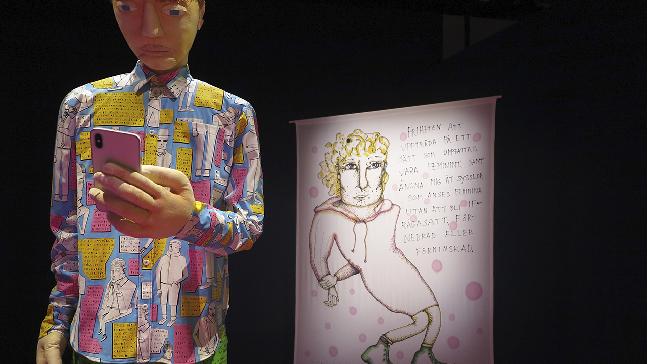 Foto som visar utställningsrummet. En skulptur av en schysst kille, klädd i kläder med citat ur utställningen.